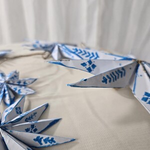 May include: A garland of white paper stars with blue painted designs. The stars are connected with twine and have a rustic, handmade look.