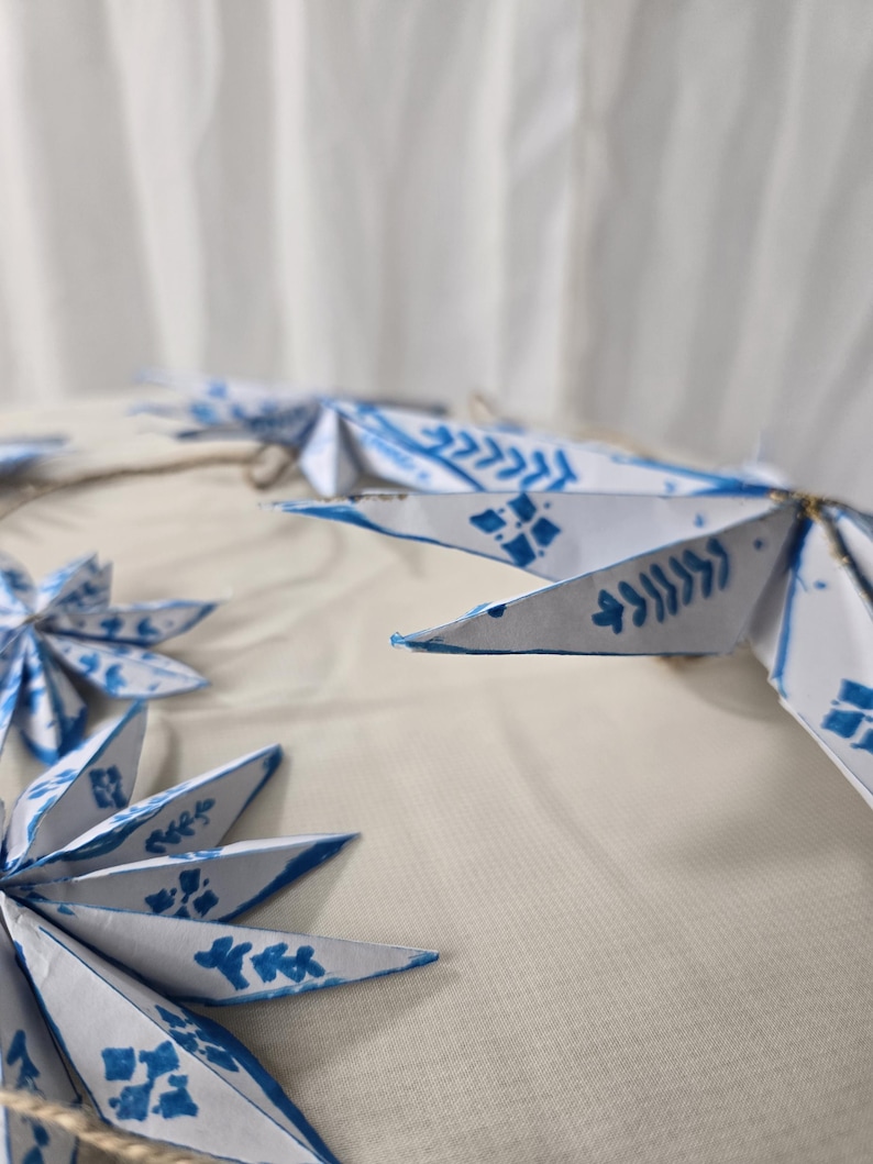 May include: A garland of white paper stars with blue painted designs. The stars are connected with twine and have a rustic, handmade look.