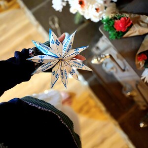 May include: A white paper star with blue and white patterned details, held in a hand. The star is folded into a three-dimensional shape.