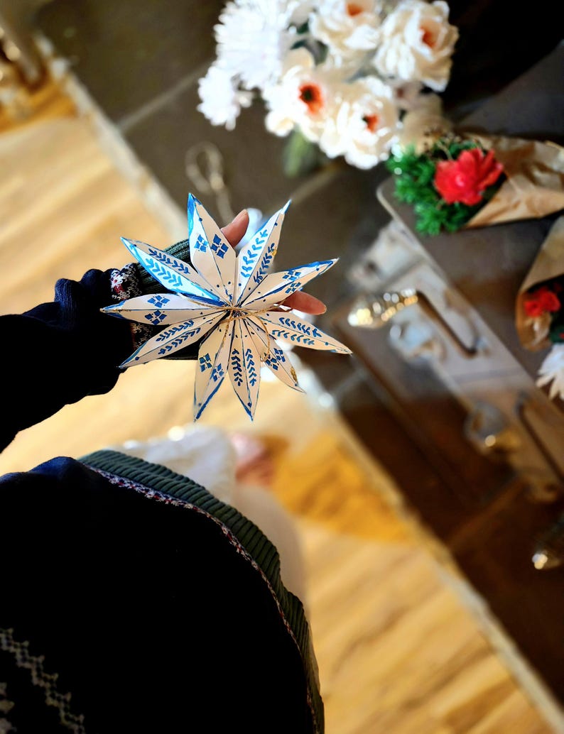 May include: A white paper star with blue and white patterned details, held in a hand. The star is folded into a three-dimensional shape.