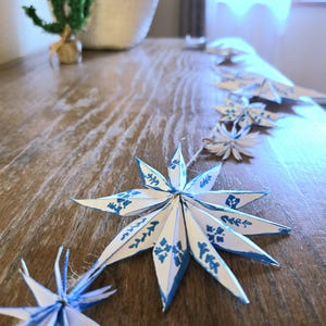 May include: A string of white paper snowflakes with blue accents hangs on a wooden surface. The snowflakes are folded into a star shape and have a blue and white pattern.