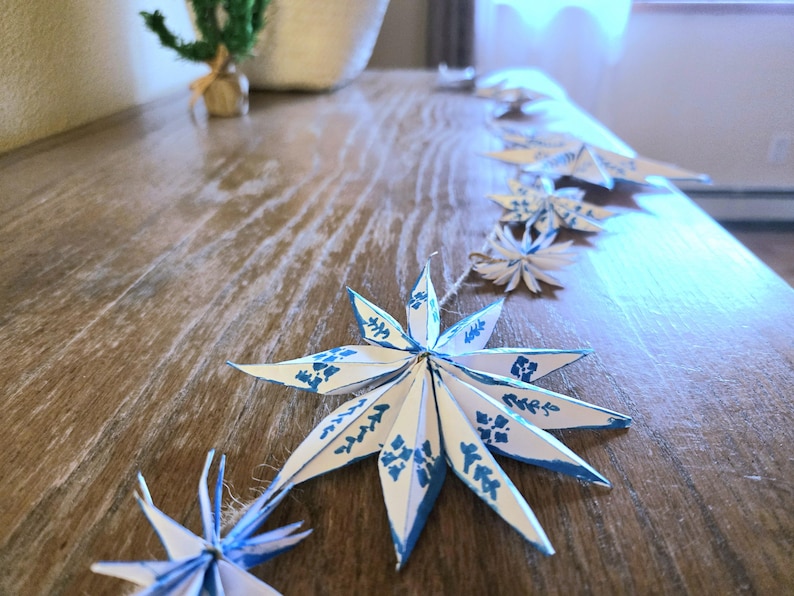 May include: A string of white paper snowflakes with blue accents hangs on a wooden surface. The snowflakes are folded into a star shape and have a blue and white pattern.