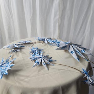 May include: A string of blue and white paper stars with a white background. The stars are connected by a twine string.