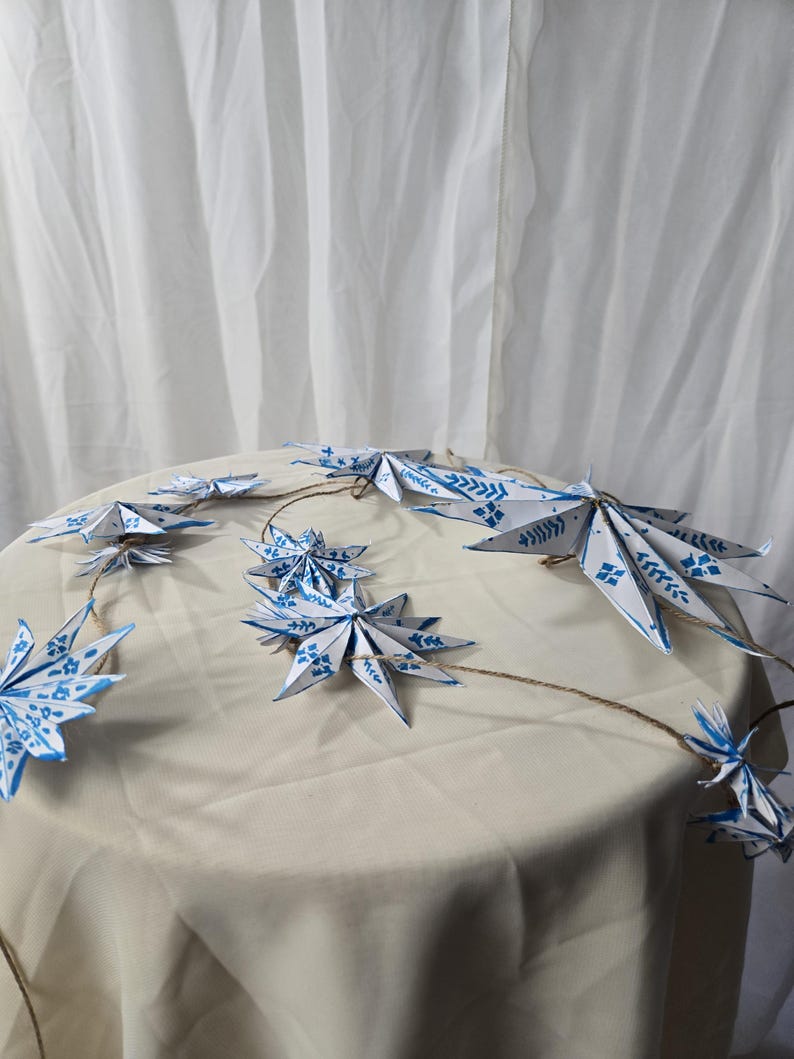 May include: A string of blue and white paper stars with a white background. The stars are connected by a twine string.