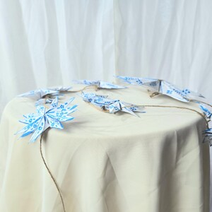 May include: A string of blue and white origami stars hanging from a table covered in a white tablecloth. The stars are connected by twine.