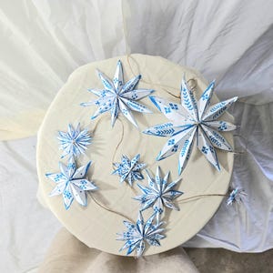 May include: A garland of blue and white paper stars with a white and blue patterned design. The stars are hanging from a string and are arranged in a circle on a white surface.
