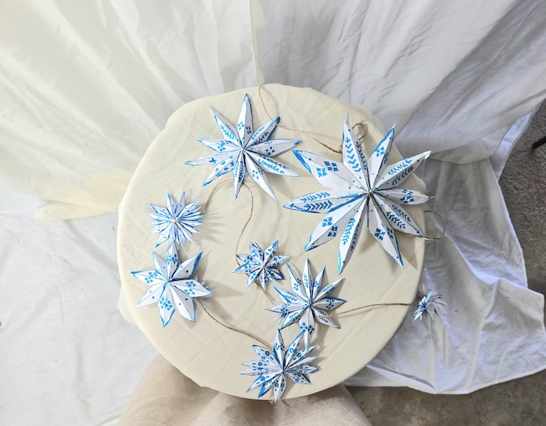 May include: A garland of blue and white paper stars with a white and blue patterned design. The stars are hanging from a string and are arranged in a circle on a white surface.