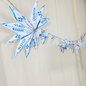 May include: A string of white paper snowflakes with blue painted designs hangs against a white background. The snowflakes are connected with twine.