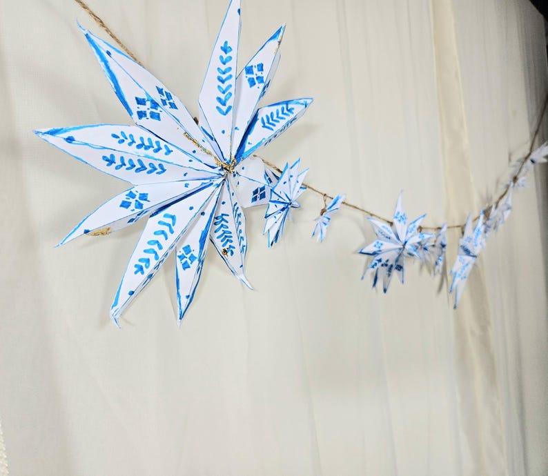 May include: A string of white paper snowflakes with blue painted designs hangs against a white background. The snowflakes are connected with twine.