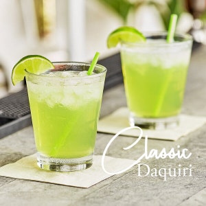 May include: Two green cocktails with ice and lime wedges in clear glasses with green straws on a white napkin. The text "Classic Daiquiri" is written on the napkin.