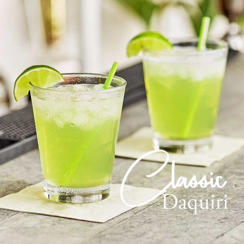 May include: Two green cocktails with ice and lime wedges in clear glasses with green straws on a white napkin. The text "Classic Daiquiri" is written on the napkin.