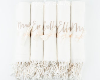 Könnte beinhalten: Fünf weiße Decken mit Fransen sind aufgerollt und mit einem Band gebunden. Jede Decke hat einen Namen in goldener Schrift gestickt: "Mad", "Em", "Holly", "Ellie" und "My".