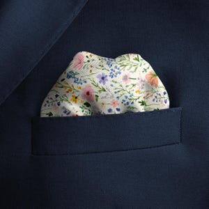 Pode incluir: Um len&ccedil;o de bolso com fundo branco apresentando um delicado padr&atilde;o floral em tons de rosa, azul, amarelo e verde, inserido no bolso do peito de um palet&oacute; azul-marinho liso com uma textura de tecido de sarja diagonal vis&iacute;vel.