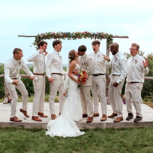 Pode incluir: Um grupo de casamento numa plataforma de madeira sob um arco floral. Um casal beija-se no centro, rodeado por seis pessoas com camisas brancas, gravatas e cal&ccedil;as bege, reagindo com entusiasmo e gestos de celebra&ccedil;&atilde;o.