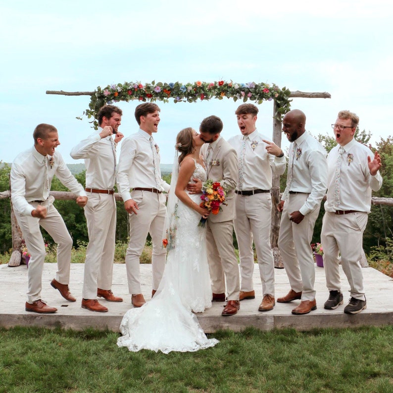 Pode incluir: Um grupo de casamento numa plataforma de madeira sob um arco floral. Um casal beija-se no centro, rodeado por seis pessoas com camisas brancas, gravatas e cal&ccedil;as bege, reagindo com entusiasmo e gestos de celebra&ccedil;&atilde;o.