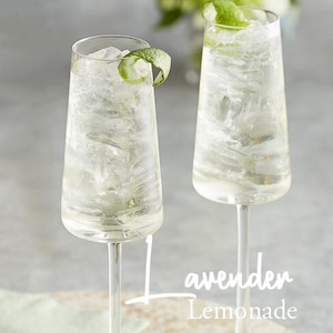 May include: Two tall, clear glasses filled with a light, refreshing beverage. The glasses are decorated with a lime wedge and ice cubes. The text "Lavender Lemonade" is written in white on a light brown surface.