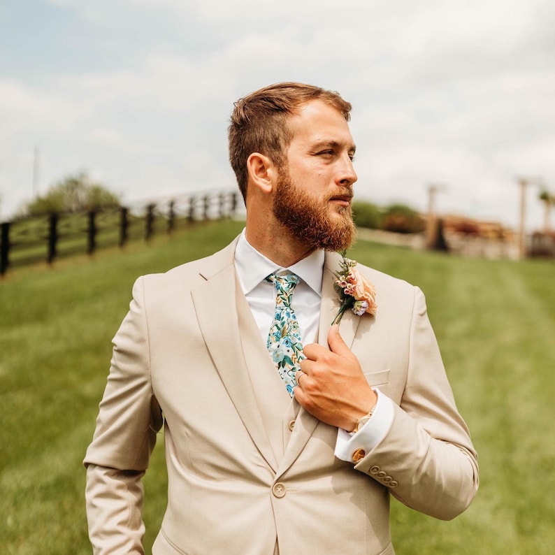 Pode incluir: Um fato de tr&ecirc;s pe&ccedil;as bege composto por uma camisa branca e uma gravata com padr&atilde;o floral azul e verde. Uma pequena flor de lapela est&aacute; presa na lapela esquerda do casaco. O conjunto inclui um colete abotoado e um blazer ajustado.