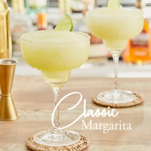 May include: Two classic margaritas in clear glass margarita glasses with a lime wedge garnish. The glasses are on woven coasters on a wooden surface. The text "Classic Margarita" is written in white cursive on the surface.