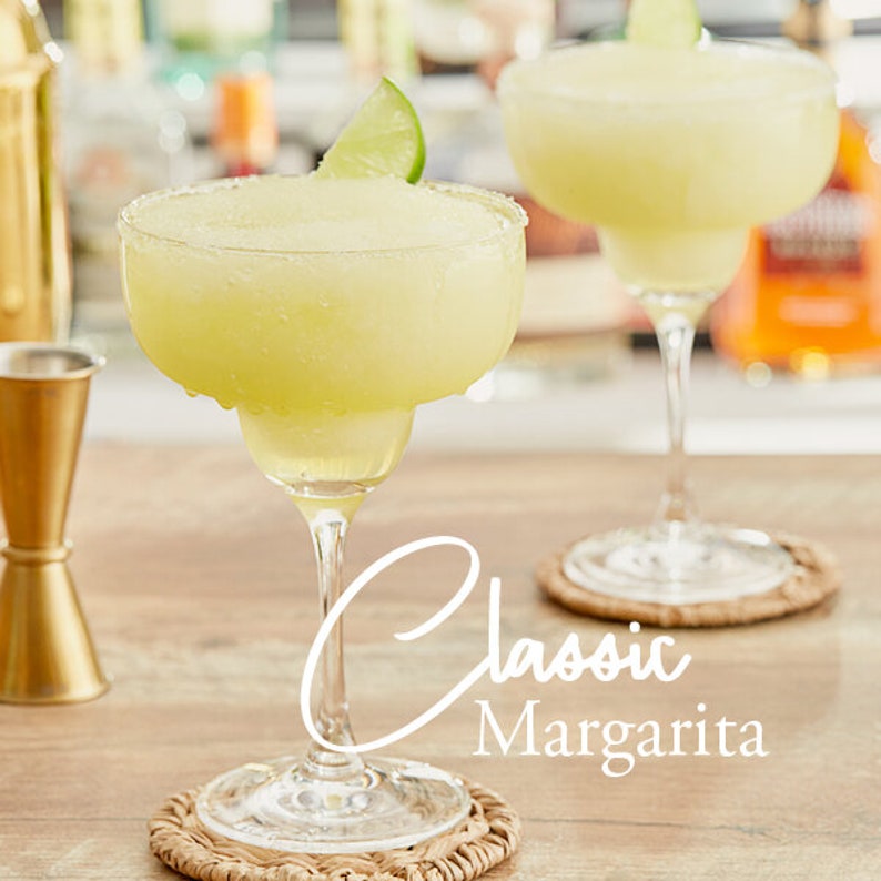May include: Two classic margaritas in clear glass margarita glasses with a lime wedge garnish. The glasses are on woven coasters on a wooden surface. The text "Classic Margarita" is written in white cursive on the surface.
