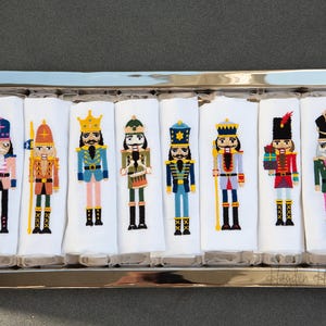 May include: Set of eight white embroidered tea towels featuring different nutcracker designs. Each towel has a different nutcracker with a unique color scheme and design. The towels are displayed in a silver tray with gold handles.