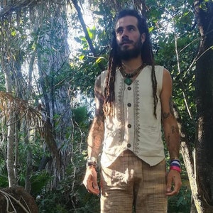 Peut inclure: Un homme avec de longues dreadlocks porte un gilet sans manches blanc avec un motif géométrique brun et blanc. Il porte également un pantalon en lin brun et un collier avec un pendentif en pierre verte.