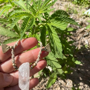 May include: A handmade necklace featuring a clear quartz crystal pendant. The necklace has a beige macrame cord with brass-colored spiral accents and small beads. The background shows green plants and sunlight.