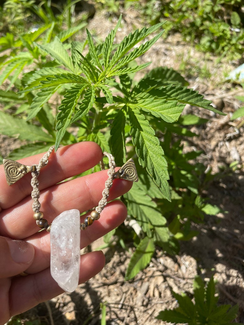 May include: A handmade necklace featuring a clear quartz crystal pendant. The necklace has a beige macrame cord with brass-colored spiral accents and small beads. The background shows green plants and sunlight.