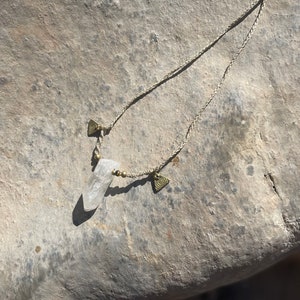 May include: A clear quartz crystal pendant necklace with a braided beige cord. The necklace features small gold-colored beads and triangular charms with spiral designs. The crystal is the focal point, suspended from the cord.