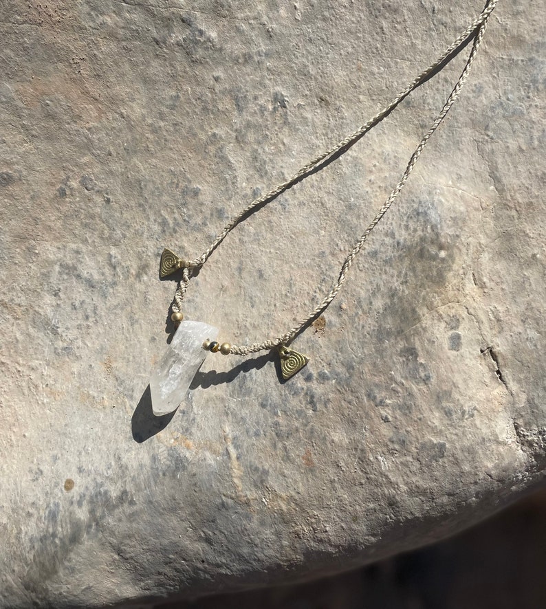 May include: A clear quartz crystal pendant necklace with a braided beige cord. The necklace features small gold-colored beads and triangular charms with spiral designs. The crystal is the focal point, suspended from the cord.
