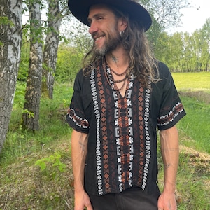 Puede incluir: Una camisa negra de manga corta con intrincados bordados marrones y blancos, combinada con pantalones oscuros. La persona lleva un sombrero marrón con una pluma y varios collares de cuentas. El fondo presenta árboles y vegetación.