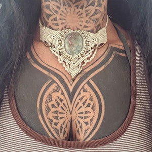 Può includere: Primo piano del petto di una persona con un grande tatuaggio di mandala in bianco e nero. La persona indossa un choker in macramé con un grande ciondolo di pietra verde.