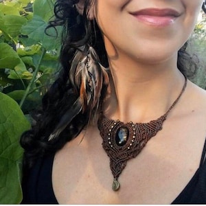 May include: A brown macrame necklace with a dark gemstone pendant. The necklace is worn by a person with long dark hair and a feather earring.