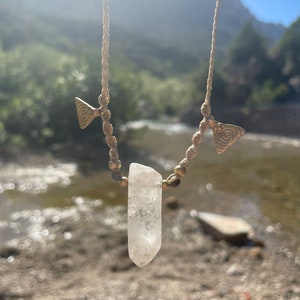 May include: A handmade necklace featuring a clear quartz crystal pendant. The necklace has a beige cord with small gold-colored beads and decorative gold-colored metal accents. The background shows a natural outdoor setting with a river and mountains.