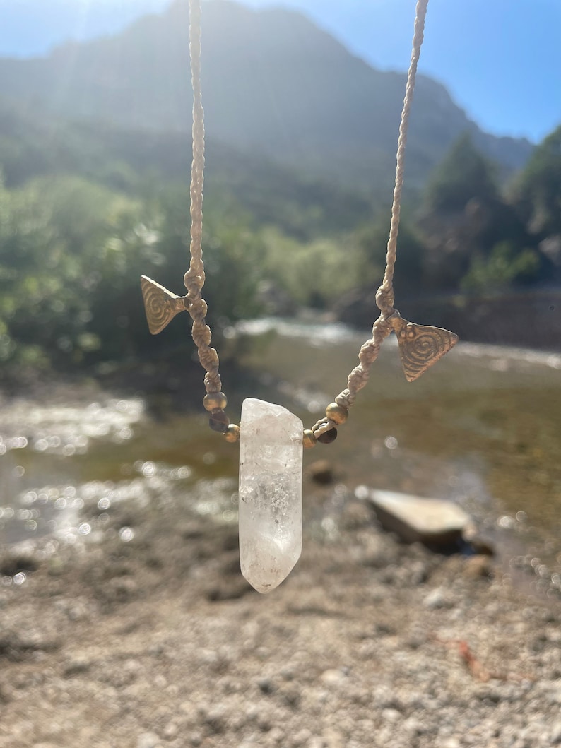 May include: A handmade necklace featuring a clear quartz crystal pendant. The necklace has a beige cord with small gold-colored beads and decorative gold-colored metal accents. The background shows a natural outdoor setting with a river and mountains.