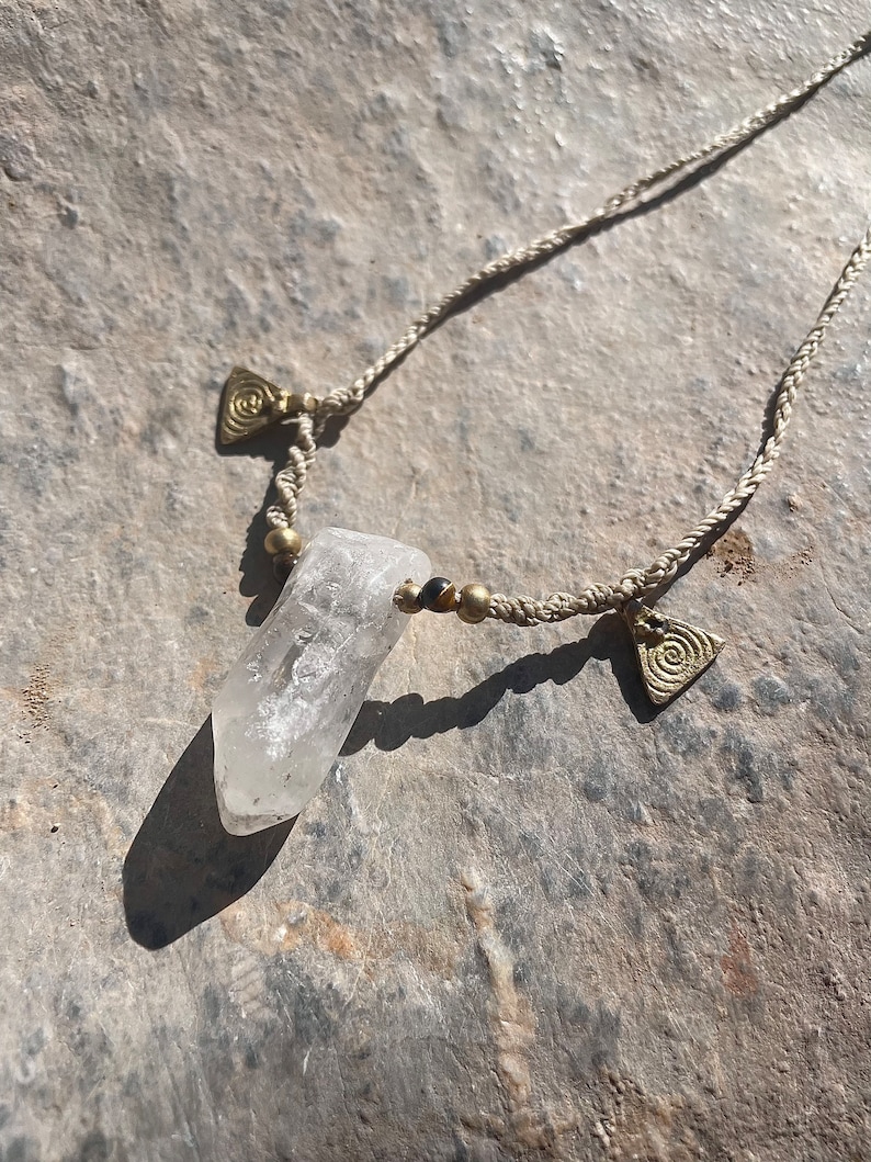 May include: A clear quartz crystal pendant necklace. The crystal is suspended from a beige woven cord, accented with brass beads and triangular charms. The necklace is displayed on a textured, gray surface.