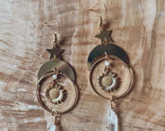 May include: Gold-toned celestial earrings featuring a star, crescent moon, and sunflower design. Each earring has a clear crystal pendant. The earrings are displayed against a wooden background.