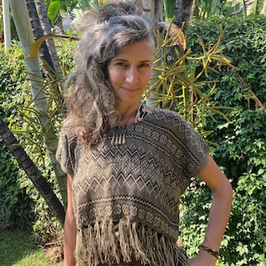 Pode incluir: Uma mulher vestindo um top marrom e branco com franjas. O top tem um design geométrico e é feito de um tecido macio e tecido. A mulher está em pé em frente a um fundo verde exuberante.