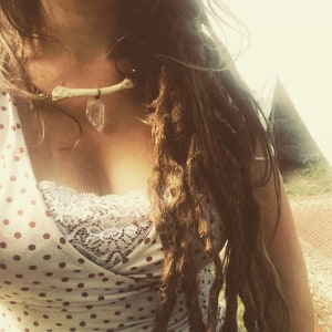 Könnte beinhalten: Eine Frau mit langen braunen Dreadlocks trägt ein weißes gepunktetes Kleid mit Spitzenbesatz und eine Halskette aus Knochen und Kristall.