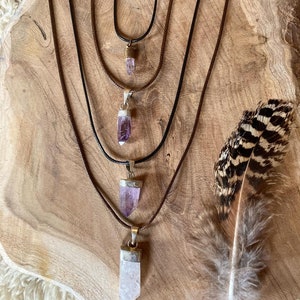 Könnte beinhalten: Fünf übereinanderliegende Halsketten mit Amethyst-Kristallanhängern an braunen Lederbändern. Die Halsketten sind in abgestufter Größe angeordnet, wobei die längste Halskette oben und die kürzeste Halskette unten liegt. Die Halsketten werden auf einer Holzoberfläche mit einer braunen Feder präsentiert.