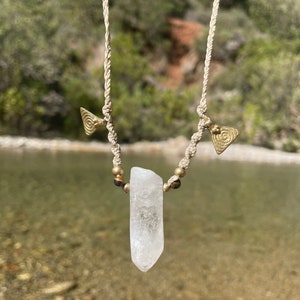May include: A handmade necklace featuring a clear quartz crystal pendant. The pendant is suspended from a beige macrame cord, accented with brass-colored beads and triangular charms. The background shows a blurred natural setting with water and greenery.