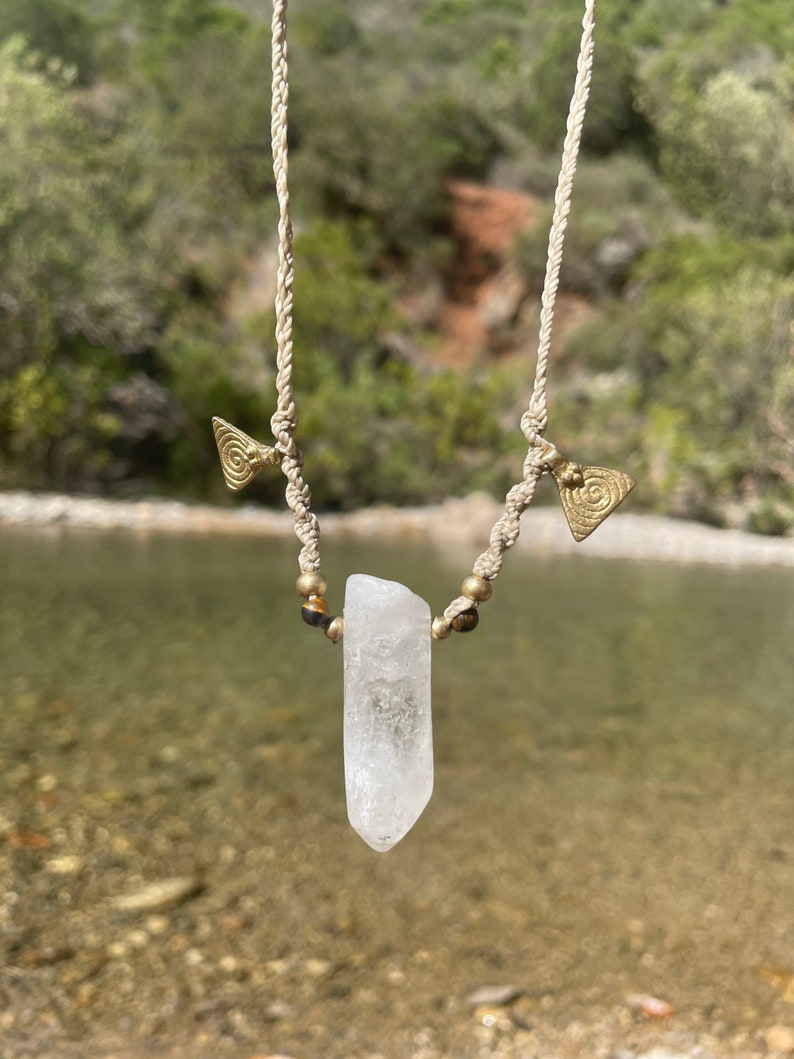 May include: A handmade necklace featuring a clear quartz crystal pendant. The pendant is suspended from a beige macrame cord, accented with brass-colored beads and triangular charms. The background shows a blurred natural setting with water and greenery.