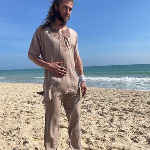 Könnte beinhalten: Eine Person trägt ein locker sitzendes, langärmeliges Hemd in hellem Braun mit Stickereien am Ausschnitt. Das Hemd wird mit einer passenden Hose kombiniert. Die Person steht an einem Sandstrand.