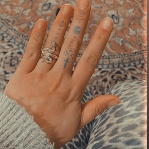 Könnte beinhalten: Ein goldener Schlangenring an einem Finger mit mehreren kleinen Tattoos, darunter ein Kreuz und ein Halbmond.