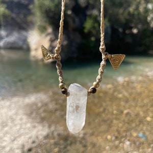 May include: A handmade necklace featuring a clear quartz crystal pendant. The pendant is suspended from a beige macrame cord, accented with small brass beads and triangular brass charms. The necklace is displayed against a natural outdoor backdrop.