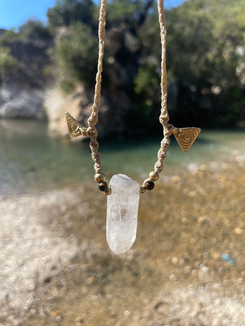 May include: A handmade necklace featuring a clear quartz crystal pendant. The pendant is suspended from a beige macrame cord, accented with small brass beads and triangular brass charms. The necklace is displayed against a natural outdoor backdrop.