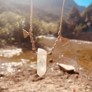 May include: A handmade necklace featuring a clear quartz crystal pendant. The necklace has a beige cord with brass-colored triangle charms and small beads. The background shows a river and mountains under a bright sky.