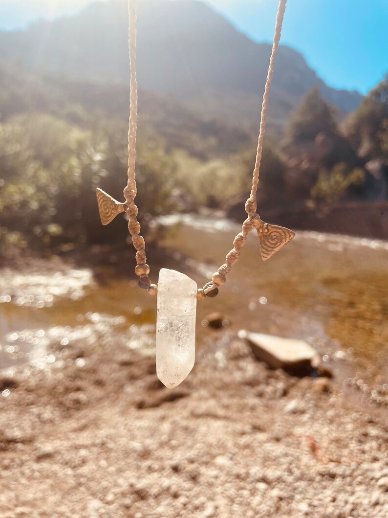 May include: A handmade necklace featuring a clear quartz crystal pendant. The necklace has a beige cord with brass-colored triangle charms and small beads. The background shows a river and mountains under a bright sky.