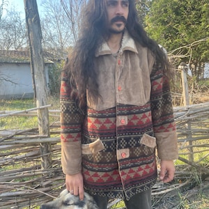May include: A man with long dark hair and a mustache is wearing a brown suede and wool jacket with a geometric pattern. He is standing next to a dog with black and white fur.