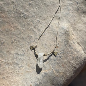 May include: A handmade necklace featuring a clear quartz crystal pendant. The necklace has a beige cord with small gold-colored beads and decorative metal accents. The crystal is suspended in the center, creating a natural and earthy aesthetic.