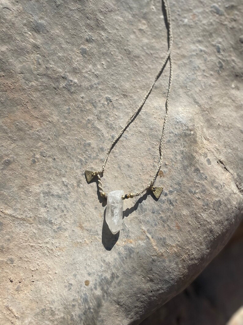 May include: A handmade necklace featuring a clear quartz crystal pendant. The necklace has a beige cord with small gold-colored beads and decorative metal accents. The crystal is suspended in the center, creating a natural and earthy aesthetic.